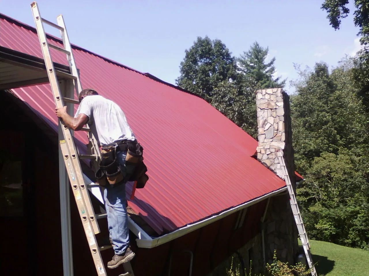 Roofer on a ladder beside a red metal roof and stone chimney — Soffit & Fascia Repair by Roofing Brothers in Byesville